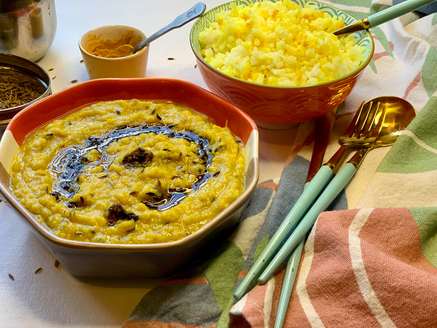 A close-up of a meal setting featuring a bowl of yellow rice and a bowl of yellow lentil dal with a drizzle of dark oil, accompanied by green-handled utensils and a patterned cloth.